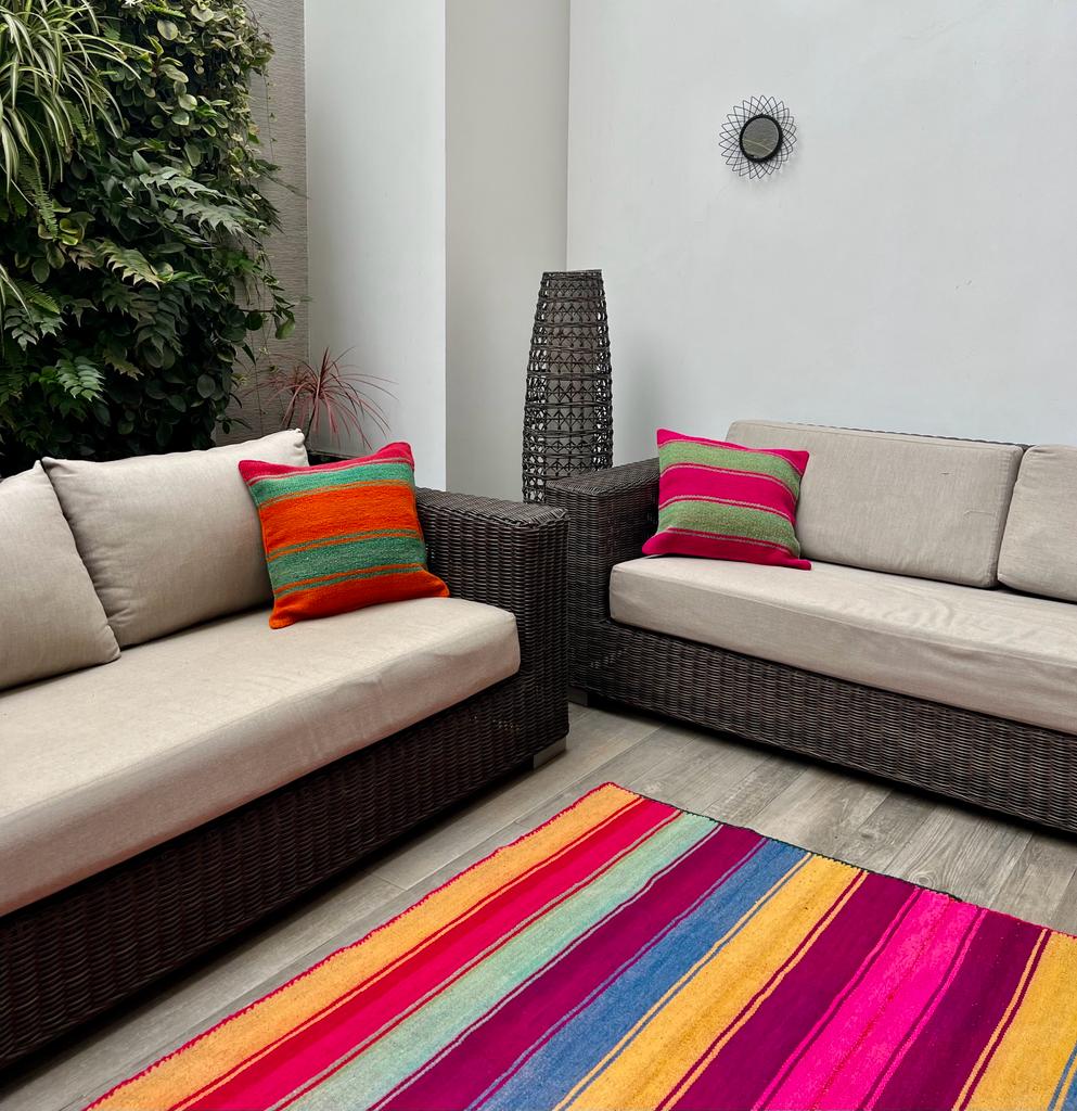 Colorful handmade Peruvian pillows and rugs displayed on an ethnic terrace. The scene features vibrant patterns and intricate designs, showcasing traditional craftsmanship. The pillows and rugs are arranged on a stone terrace, surrounded by potted plants and overlooking a picturesque view.